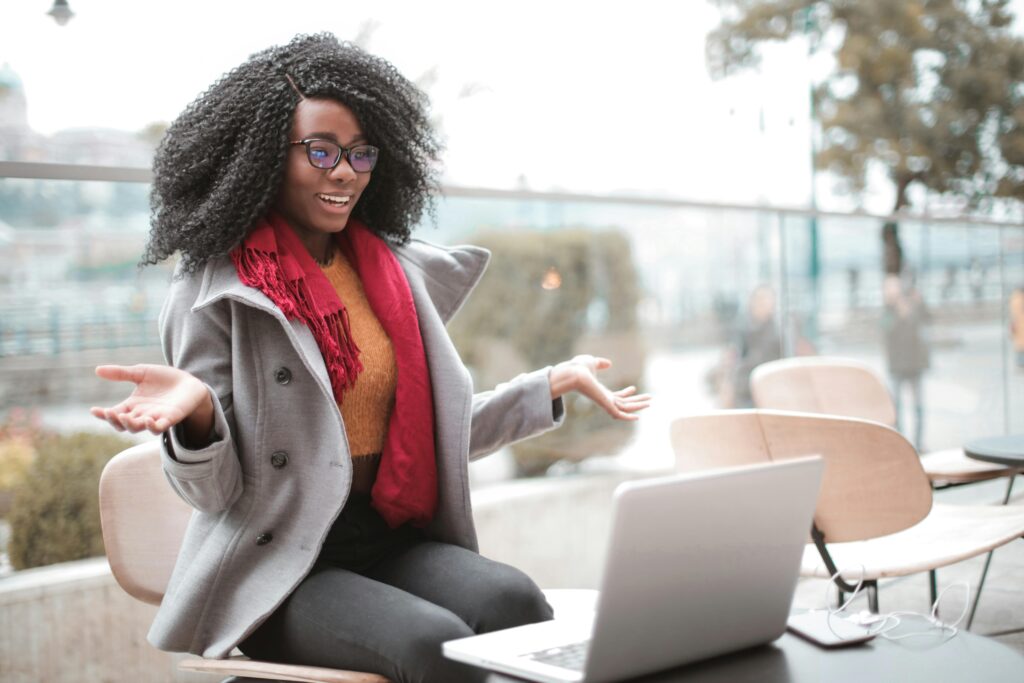 A cheerful woman with curly hair wearing glasses and a grey coat sits at an outdoor cafe table, joyfully interacting with her laptop which is on an advanced and user-friendly e-commerce website, a satisfied smile on her face suggesting a pleasant online shopping experience.